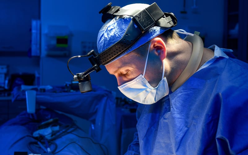 A surgeon wearing a head-mounted surgical headlight system with an LED/fiber-optic light module during a procedure