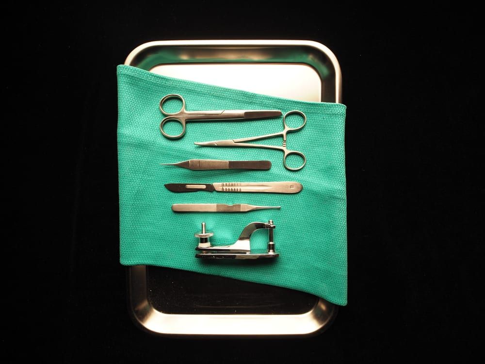 An assorted stainless-steel surgical instrument set — scissors, a needle holder, tissue forceps, and a scalpel — laid out in a stainless instrument tray
