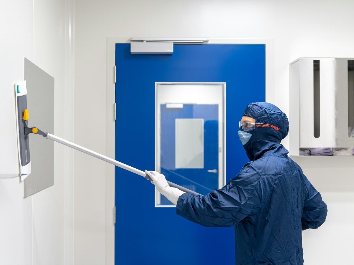 Technician cleaning a cleanroom wall panel beside a door with a vision panel