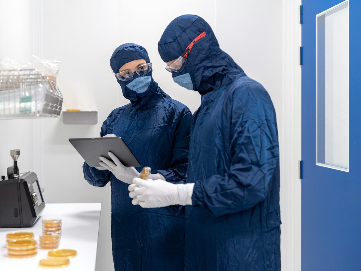 Two gowned technicians reviewing settle plates beside an air sampler